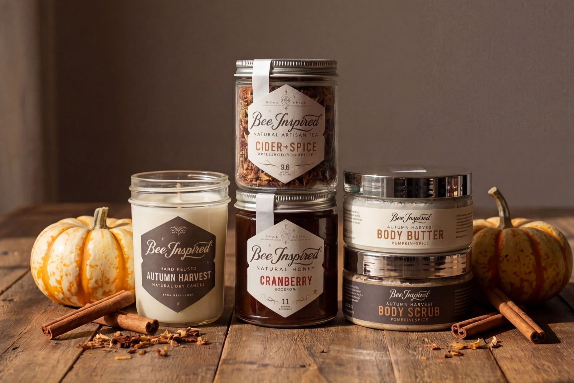 Collection of Bee Inspired products including candles and body scrubs on a wooden surface with pumpkins and cinnamon sticks.