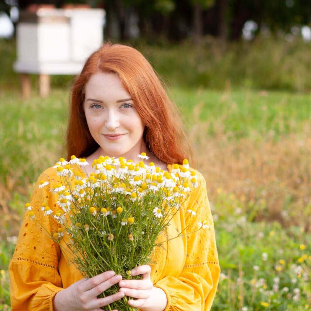 a redheaded woman in a yard, holding a bouquet of wild chamomile flowers