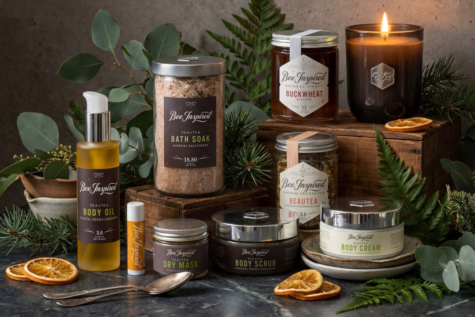 Collection of Bee Inspired Ultimate Winter Respite skincare and wellness products with candles and herbs on a dark surface.