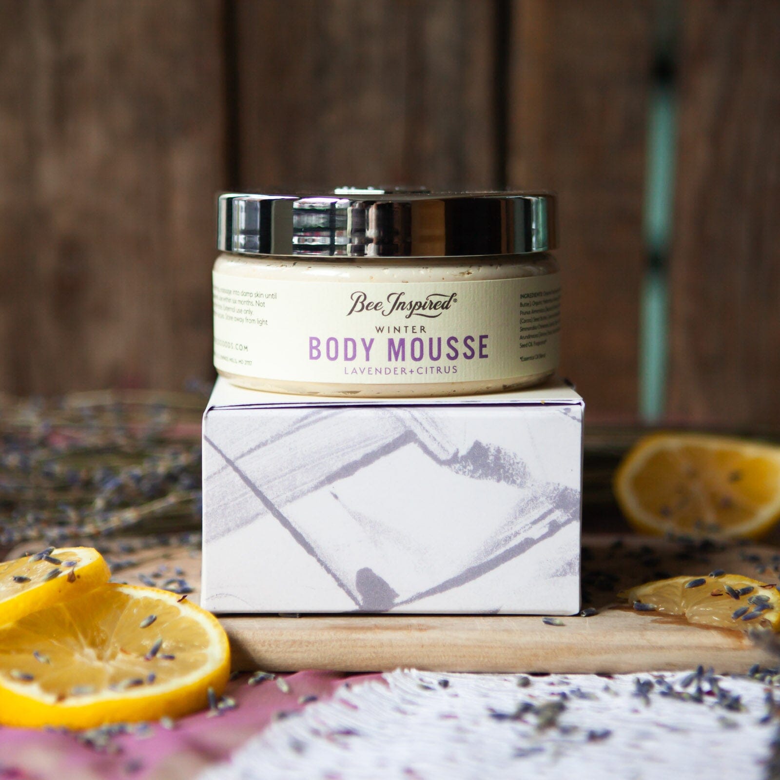 Bee Inspired Winter Body Mousse sitting on a box covered with artwork. The butter and box are sitting upon a cutting board and are surrounded by dried citrus and lavender.