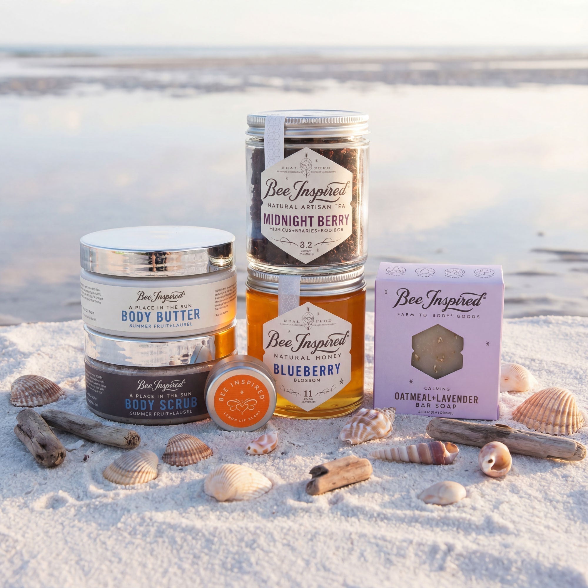 Bee Inspired product lineup including jars, a box, and a bar on a sandy beach with seashells.