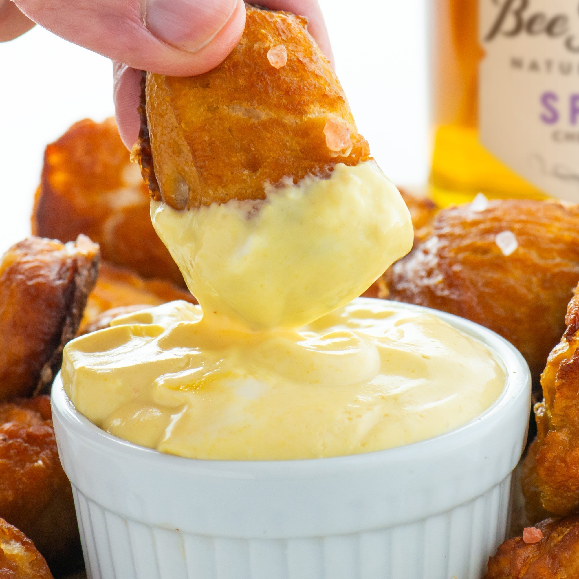 Dipping a soft pretzel into a bowl of honey mustard sauce with a background of more soft pretzels and a jar of Spring Honey.