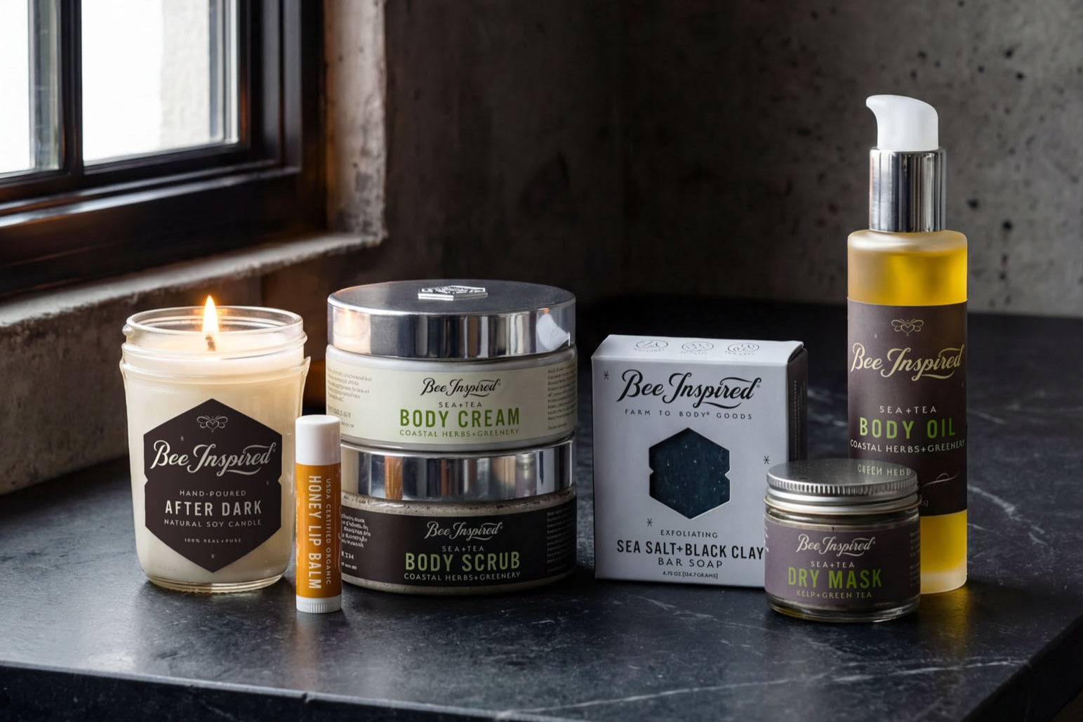 Collection of Bee Inspired skincare and grooming products from the King Bee Body Care Kit on a dark surface with a window in the background.