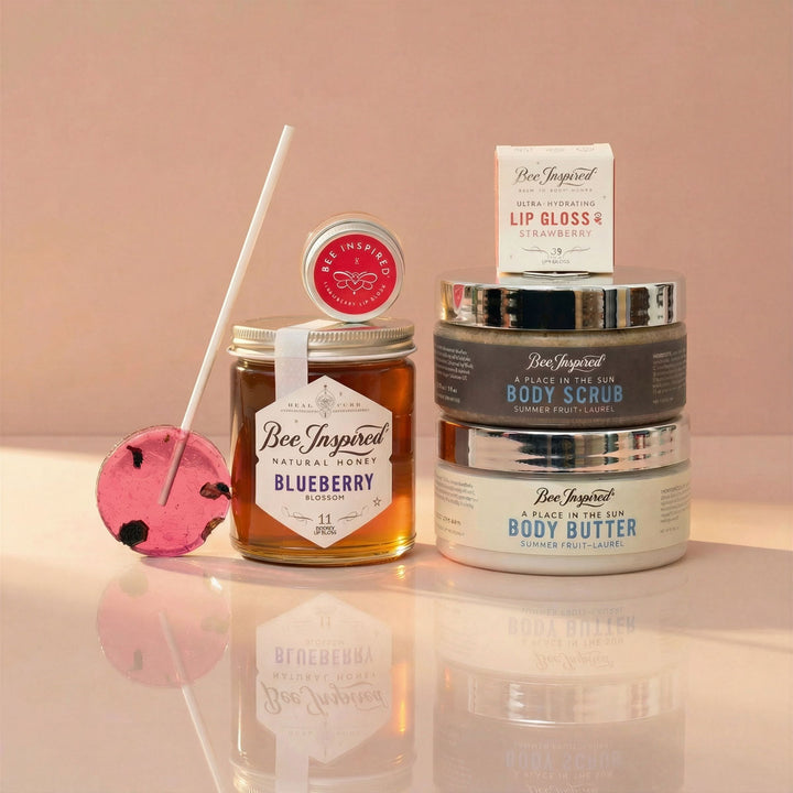 Bee Inspired skincare products including lip gloss, body scrub, and body butter on a reflective surface with a pink background.