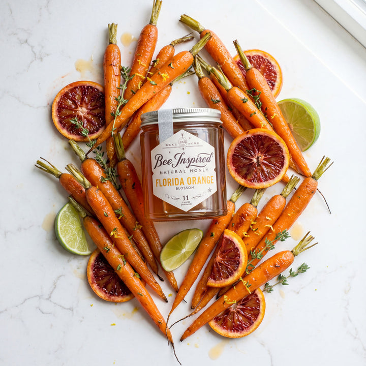 Jar of 'Bee Inspired' Florida Orange Blossom honey surrounded by carrots, oranges, and limes on a marble surface.