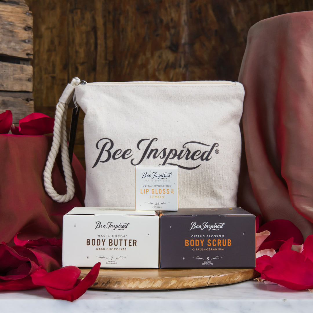 BeeInspired skincare products including a pouch, lip gloss, body butter, and body scrub on a wooden surface with red flowers.