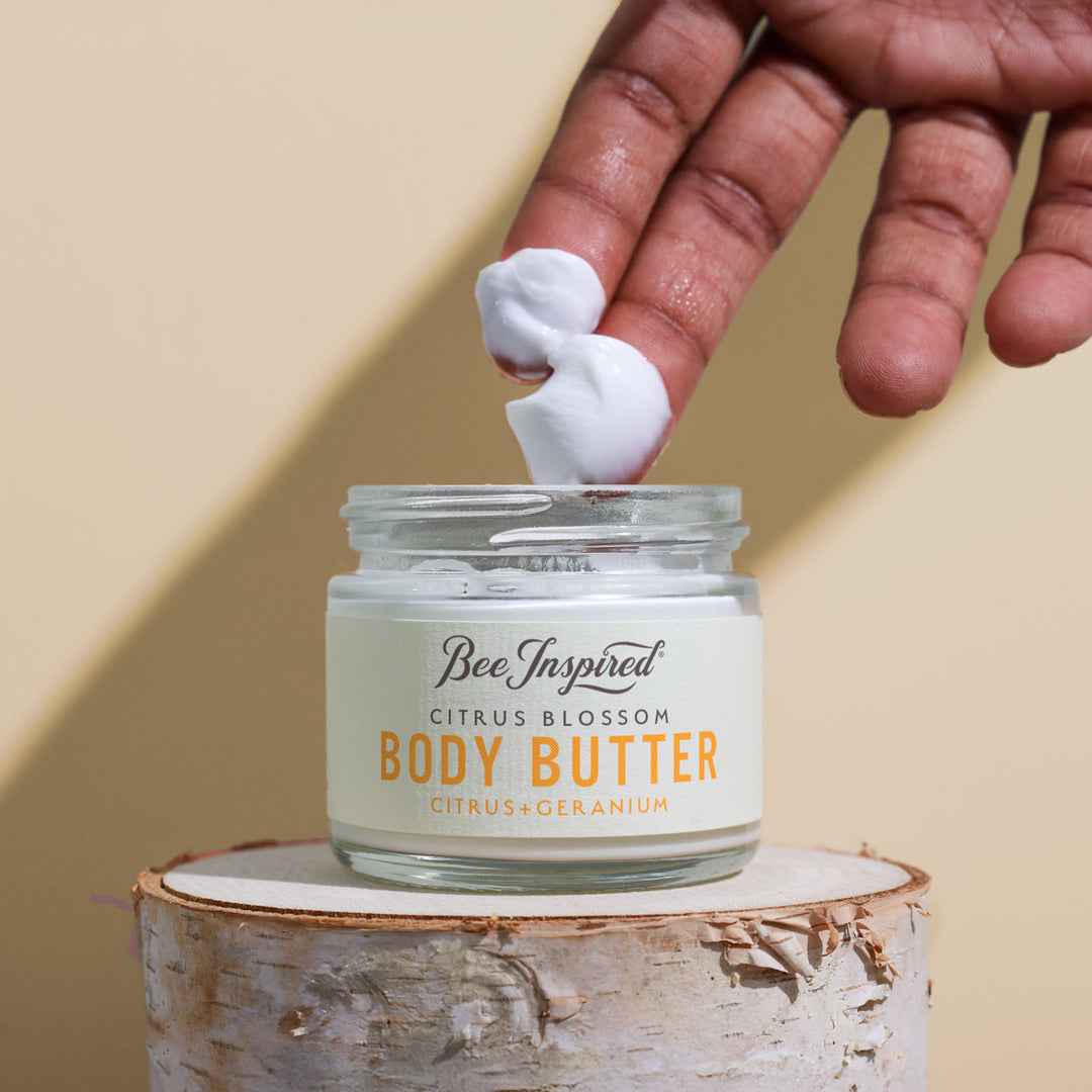 Hand applying body butter to a jar labeled 'Bee Inspired Citrus Blossom Body Butter' on a wooden block with a beige background.