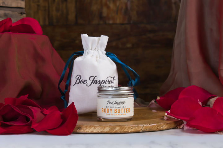 Bee Inspired body butter jar with a drawstring bag on a wooden board surrounded by red flowers.
