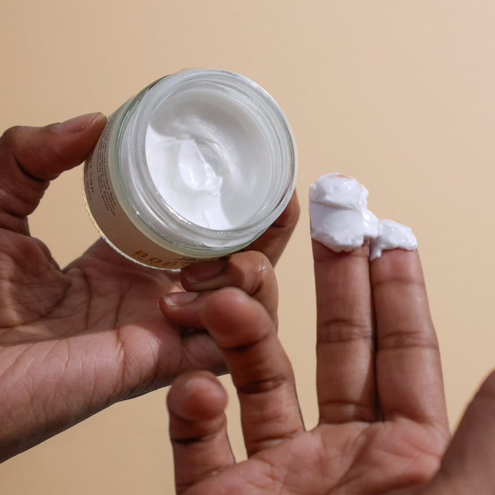Hand holding a jar of white cream with a small amount on the finger against a beige background