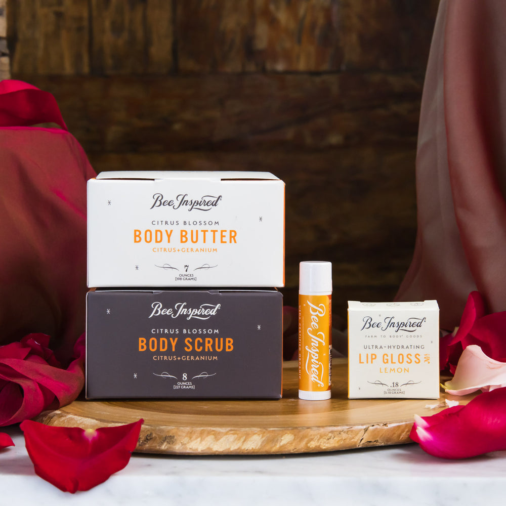 Bee-Inspired skincare products including body butter, scrub, lip gloss, and lip balm on a wooden tray with red flowers.