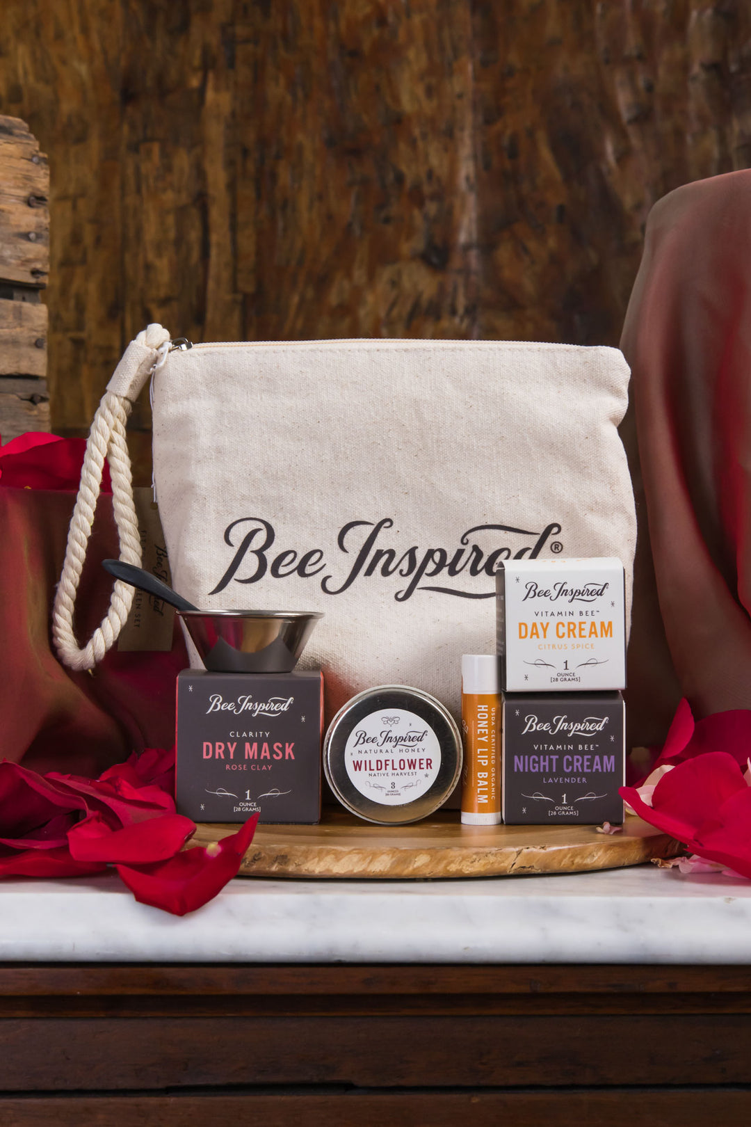 BeeInspired skincare products including a pouch, cream bottles, and containers on a wooden surface with red flowers.