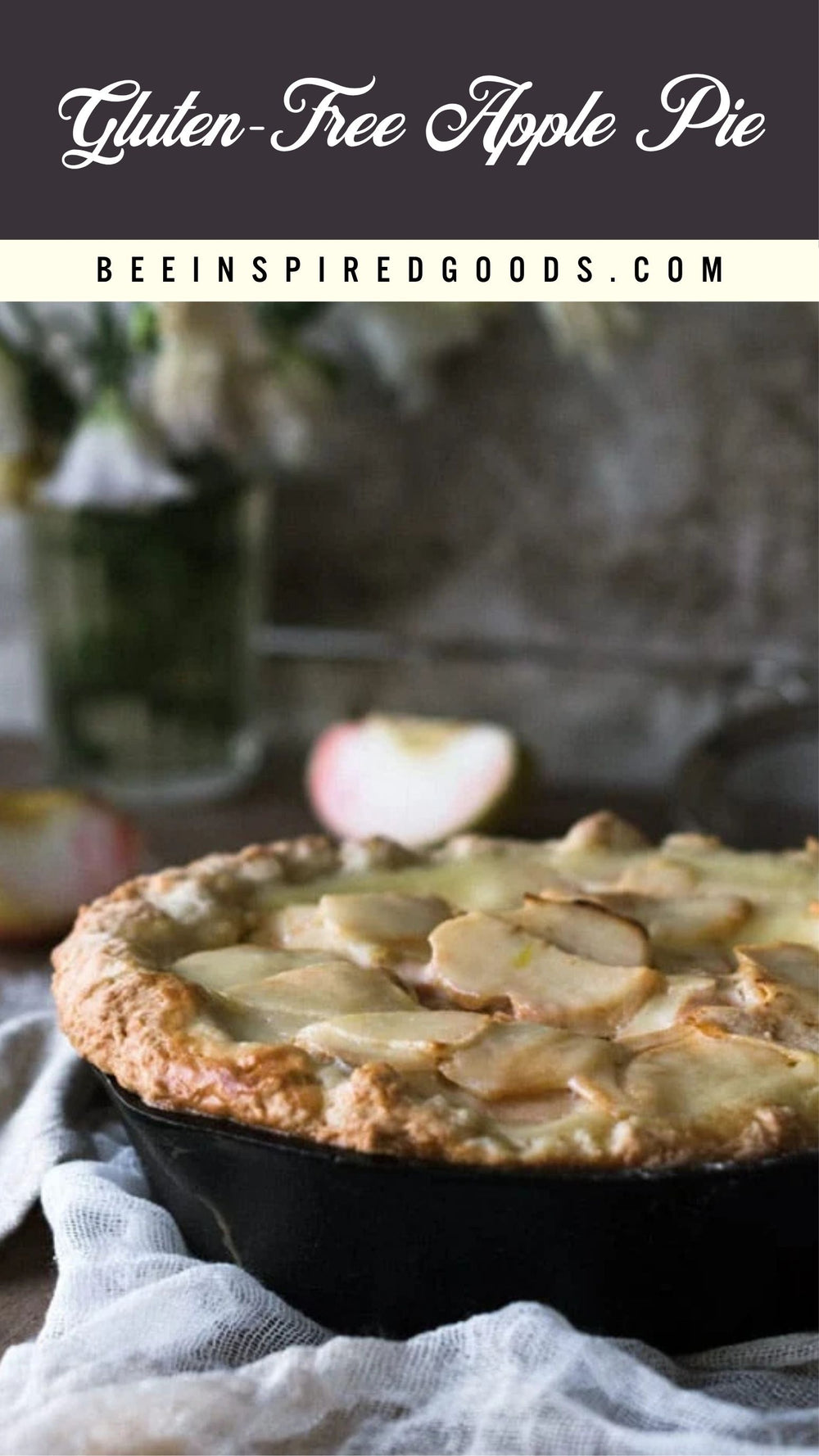 A gluten-free apple pie in a black cast iron skillet, topped with sliced apples and a golden crust, set on a white cloth with blurred apples and a vase in the background. Text at the top reads 'Gluten-Free Apple Pie' and 'BEEINSPIREDGOODS.