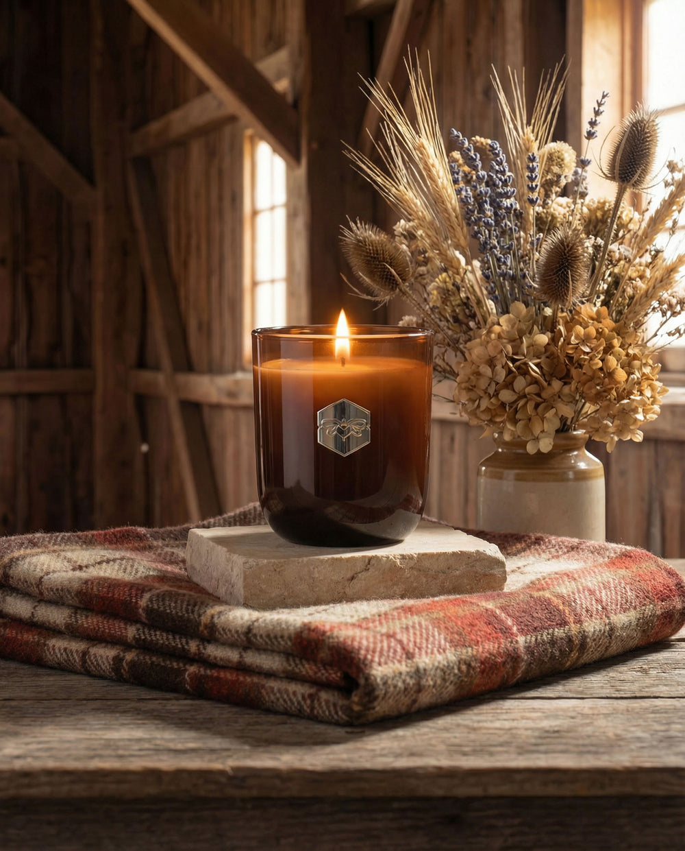 Candle with a hexagonal logo on a wooden surface with a plaid blanket and dried flowers in the background.