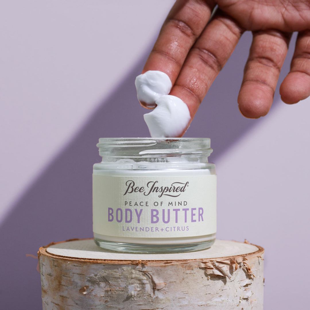 Hand applying body butter to a jar labeled 'Bee Inspired Peace of Mind Body Butter' on a wooden block with a purple background.