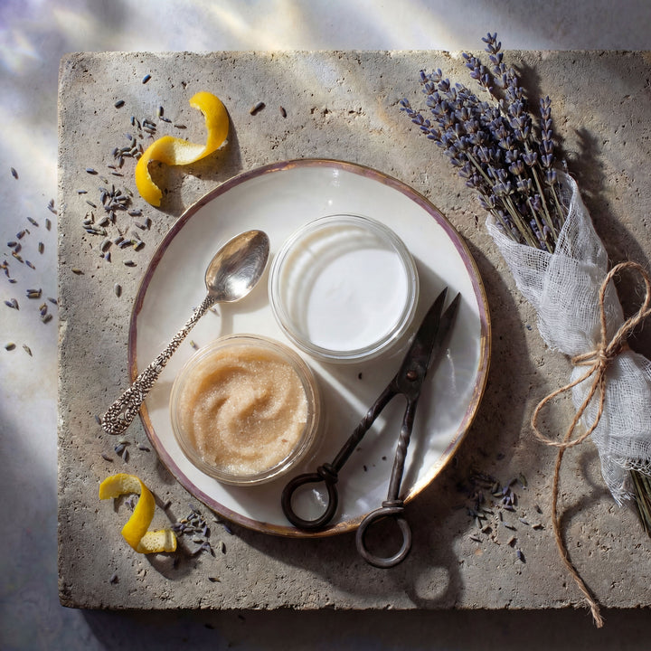 A jar of body butter and a jar of body scrub with spoons on a stone surface, surrounded by lavender flowers and petals.