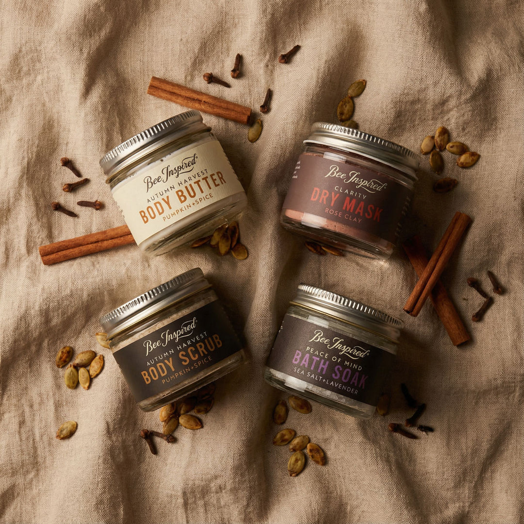 Four jars of Bee Inspired body care products on a textured fabric background with cinnamon sticks and cardamom pods.