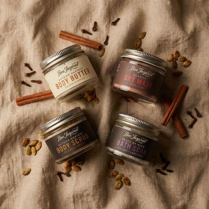 Four jars of Bee Inspired body care products on a textured fabric background with cinnamon sticks and cardamom pods.