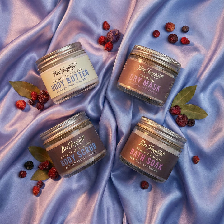 Four jars of Bee Inspired body care products on a textured purple fabric background with berries and leaves.