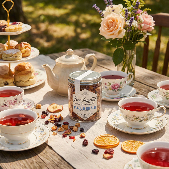 Tea party setup with tea cups, teapot, and a jar of 'Bee Inspired' place in the sun tea on a wooden table outdoors.