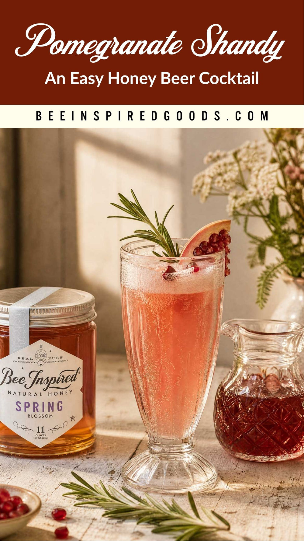 A styled cocktail scene featuring a tall glass of pinkish-orange pomegranate shandy with rosemary and pomegranate garnish, alongside a jar labeled Bee Inspired Goods Spring Blossom honey and a small pitcher of dark red liquid on a rustic table.