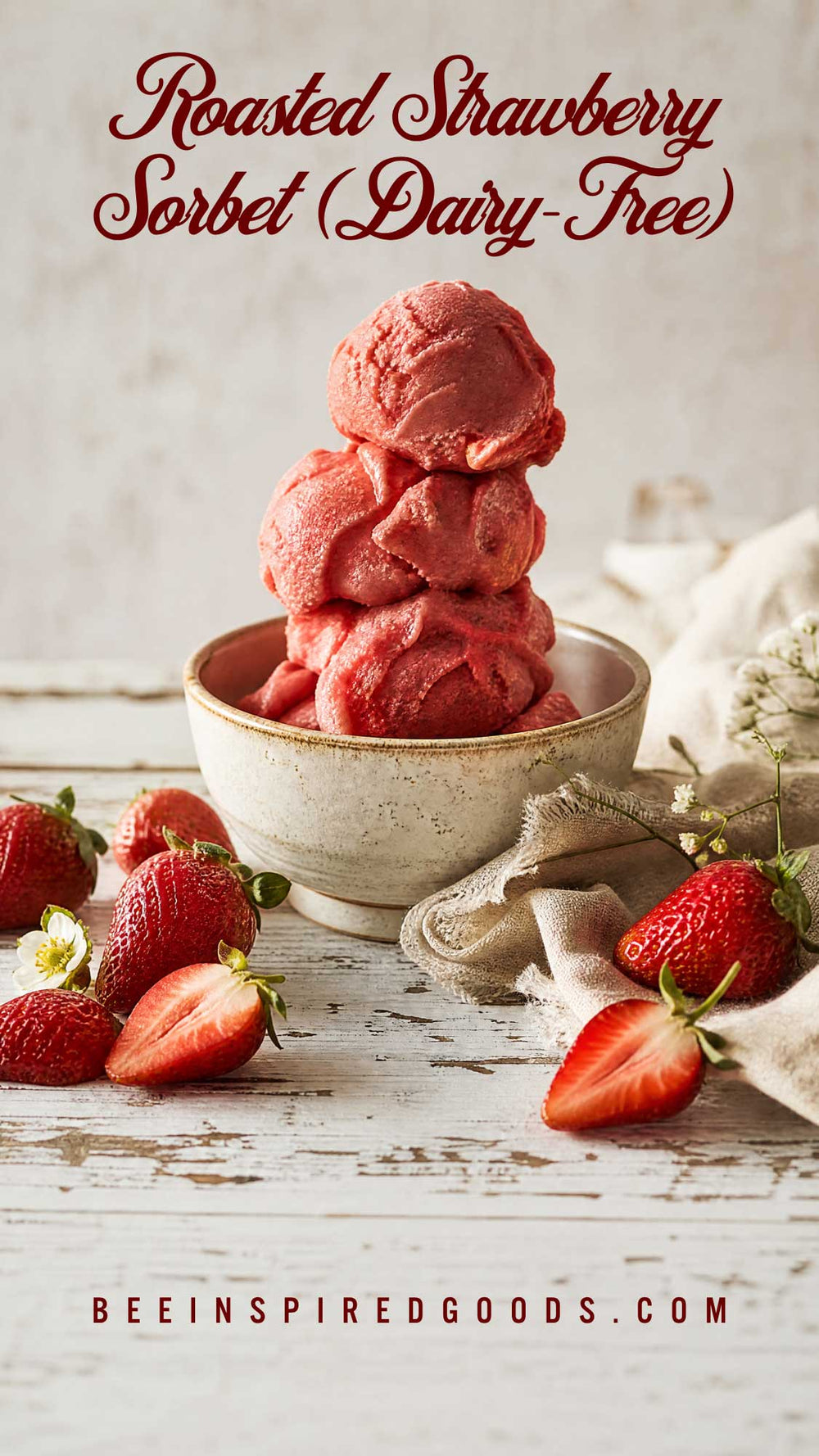 A styled food photo showing scoops of pink-red roasted strawberry sorbet in a small bowl on a rustic white wooden surface, with whole and sliced strawberries around it and text reading 'Roasted Strawberry Sorbet (Dairy-Free)' and 'BEEINSPIREDGOODS.COM'.