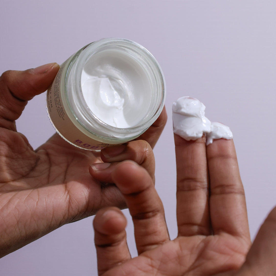 Hand holding a jar of cream with some on the finger against a light purple background
