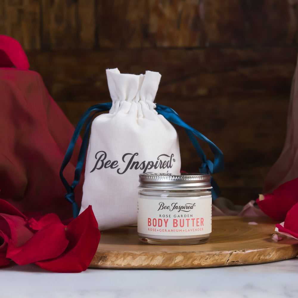 Jar of body butter with a drawstring bag labeled 'Bee Inspired' on a wooden board with red flowers.