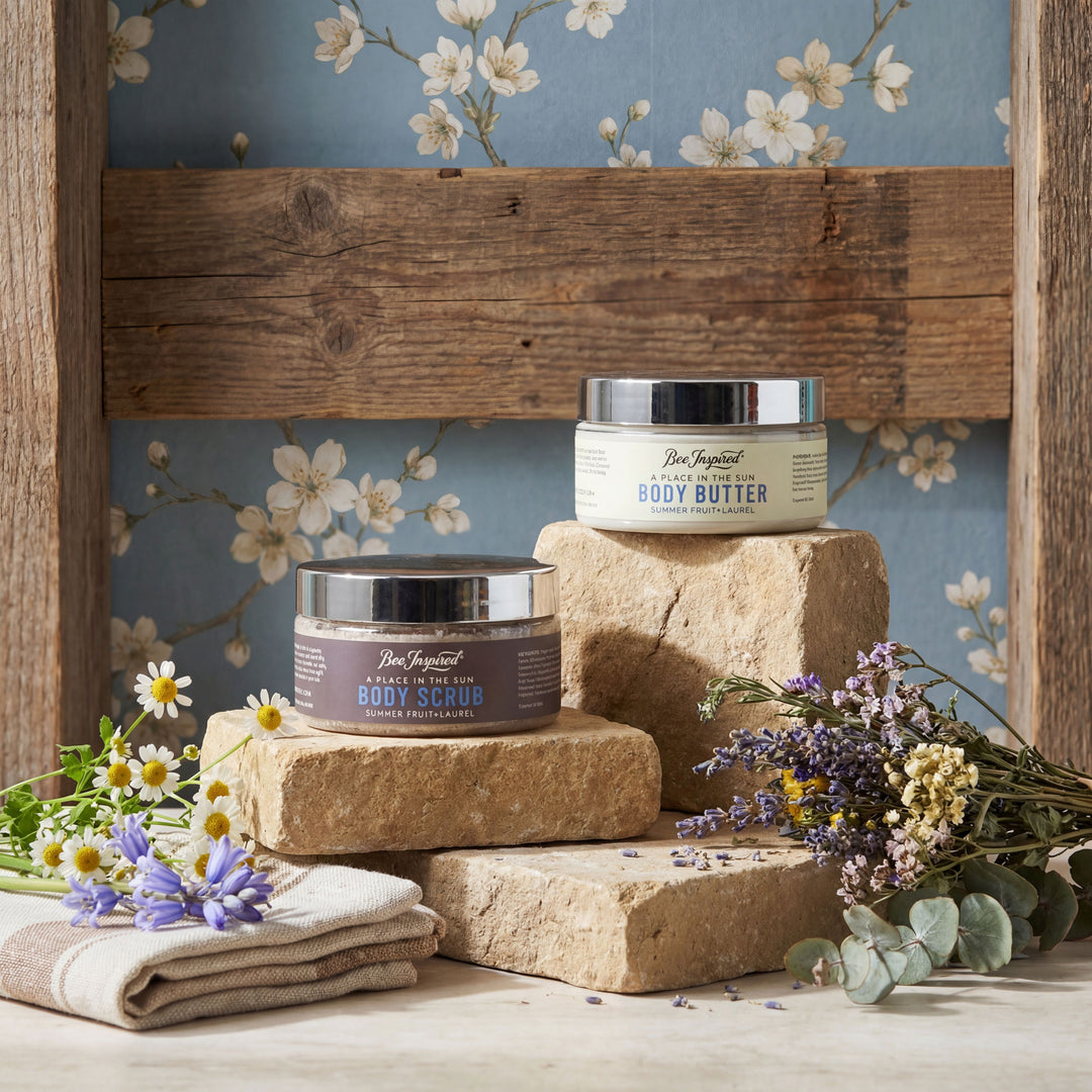 Two jars of body butter on stone blocks with flowers and a wooden shelf in the background.