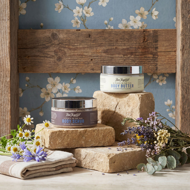 Two jars of body butter on stone blocks with flowers and a wooden shelf in the background.