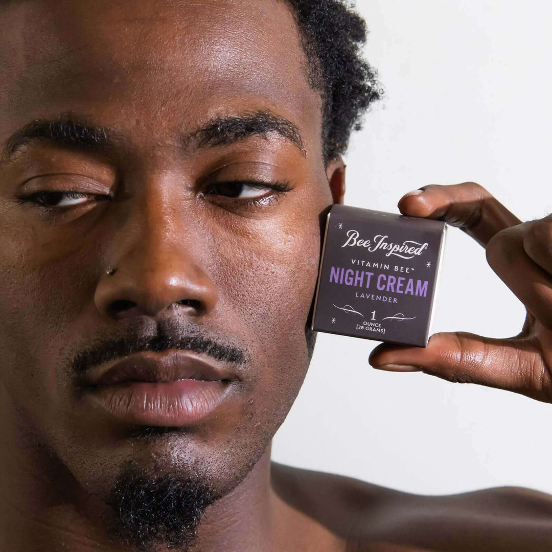 man holds Vitamin Bee lavender night cream by his face and looks away from the camera