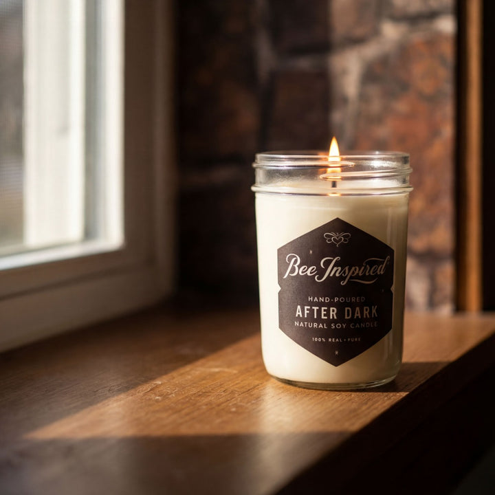 Candle in a jar labeled 'Bee Inspired After Dark' on a wooden surface with a window and stone wall in the background.