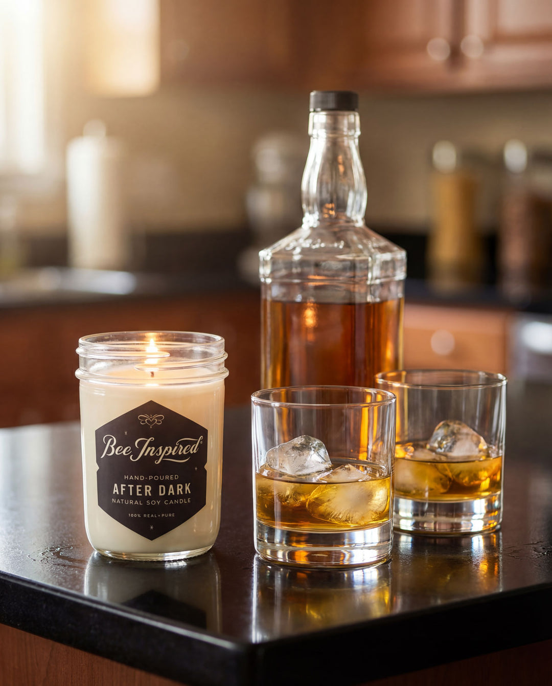 Candle labeled 'Bee Inspired After Dark' next to a bottle and two glasses of whiskey on a counter.