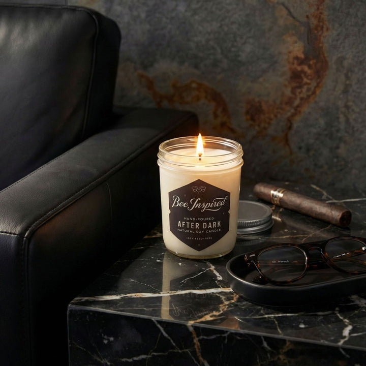 Candle labeled 'Bee Inspired After Dark' on a marble surface with a black leather chair and glasses in the background.