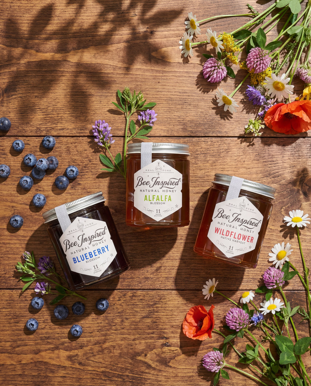 Three jars of 'Bee Inspired' honey on a wooden surface with flowers and blueberries.