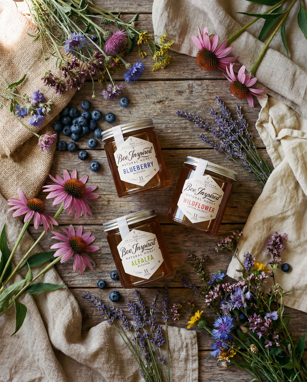 Three jars of honey labeled 'Blueberry', 'Wildflower', and 'Alfalfa' on a wooden surface with flowers and blueberries.