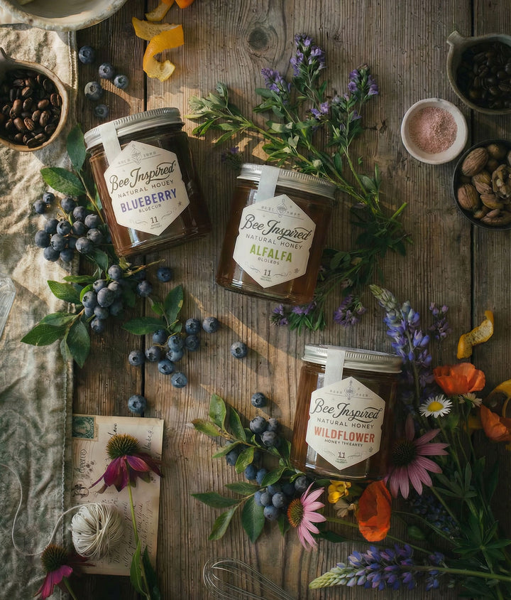 Three jars of honey labeled 'Blueberry', 'Alfalfa', and 'Wildflower' on a wooden surface with flowers and berries.