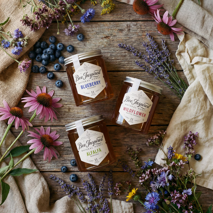 Three jars of 'Bee Inspired' honey surrounded by flowers and blueberries on a wooden surface.