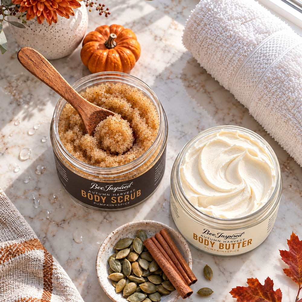 Two jars of Bee Inspired body products on a marble surface: an open jar of brown sugar scrub with a wooden spoon and a closed jar of cream-colored body butter. Decorative autumn props including a small pumpkin, cinnamon sticks, pumpkin seeds, and a rolled towel surround the products.
