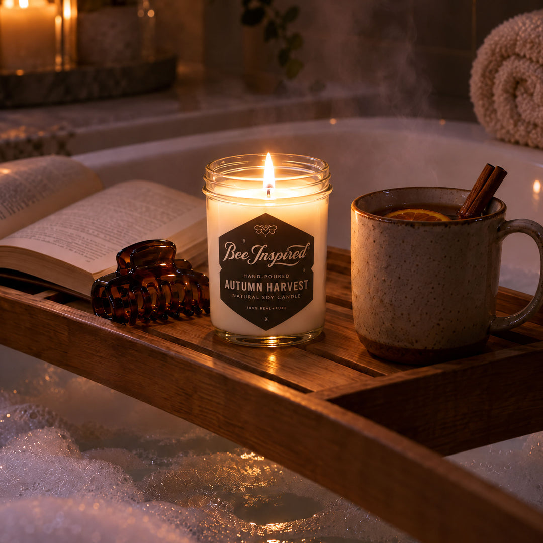 A lit candle in a clear glass jar sits on a wooden bath caddy in a hot tub, surrounded by an open book, a brown claw clip, and a speckled mug with a cinnamon stick. The candle label reads &