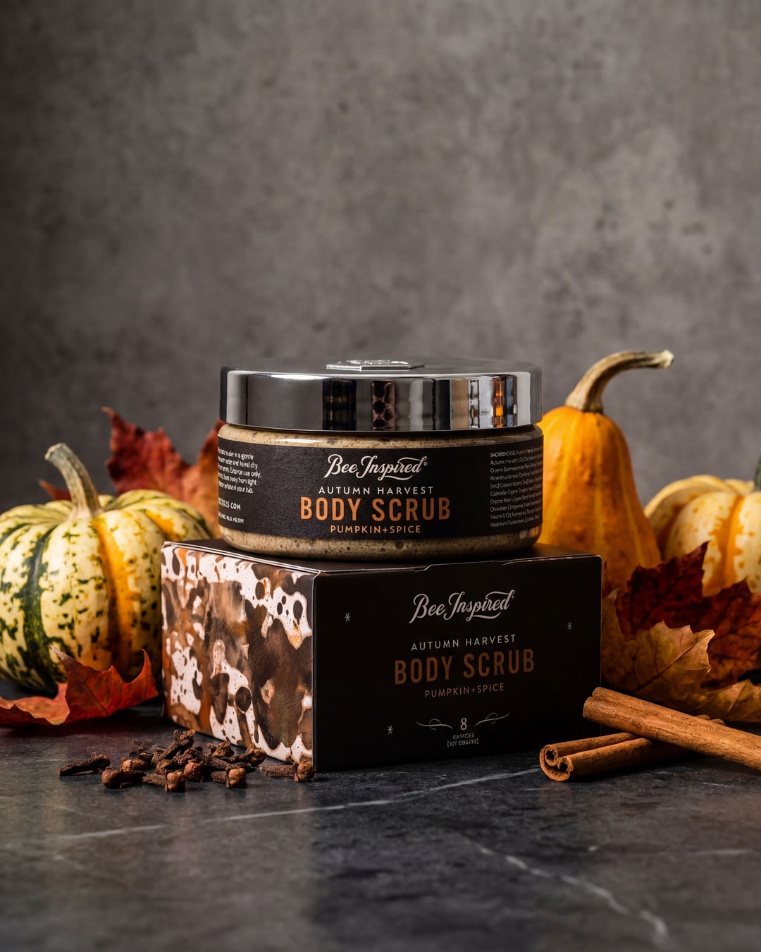Bee Inspired Body Scrub with packaging on a dark surface with pumpkins and cinnamon sticks.