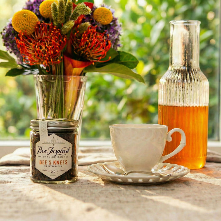 Tea cup with saucer, jar of 'Bee's Knees' tea, and bottle of honey on a linen clothe with tees in the background.
