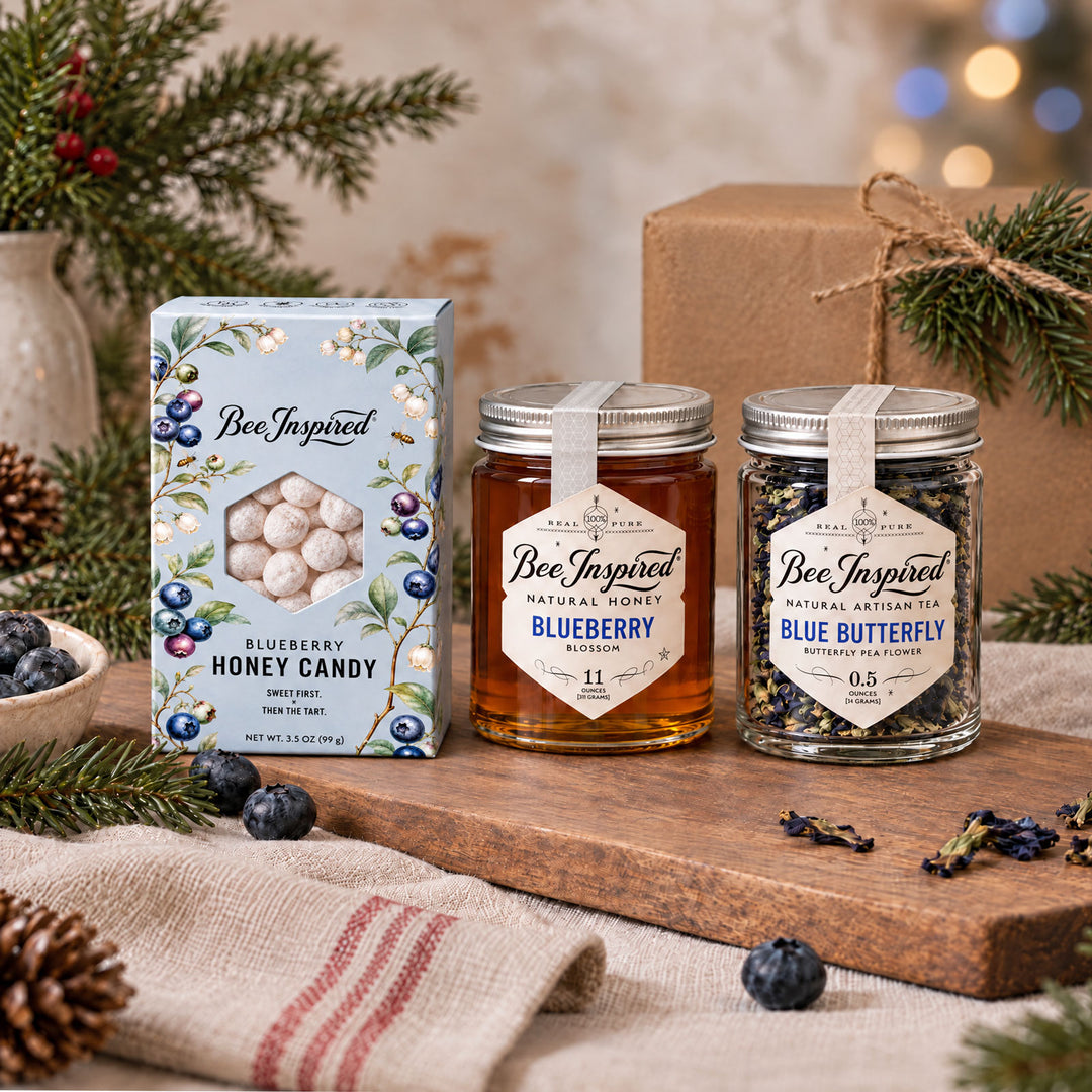 Blue Christmas gift set of blueberry honey candy, blueberry blossom honey, butterfly blue pea tea on wood surrounded by greenery and gifts and some blurry holiday lights.