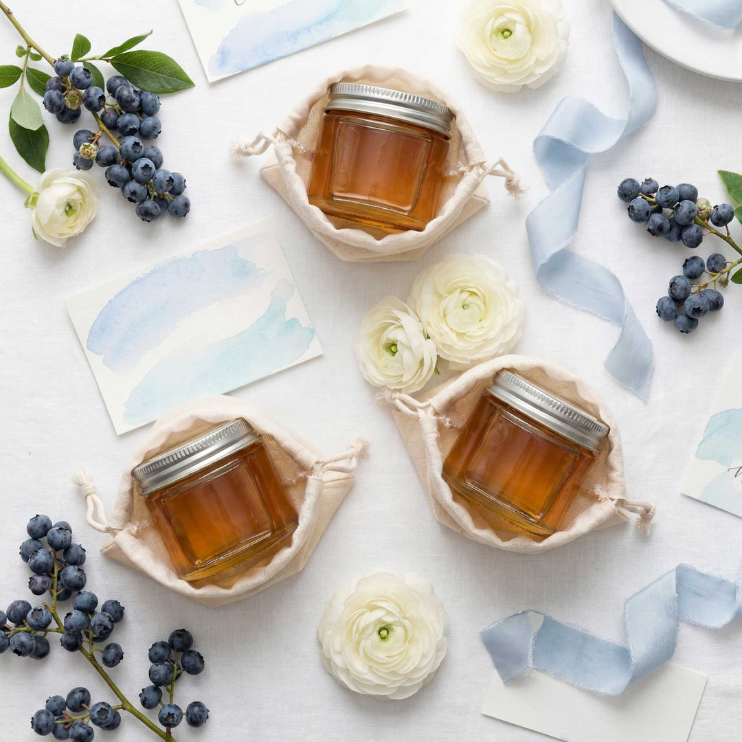 Three jars of honey on a white surface with flowers and blueberries.