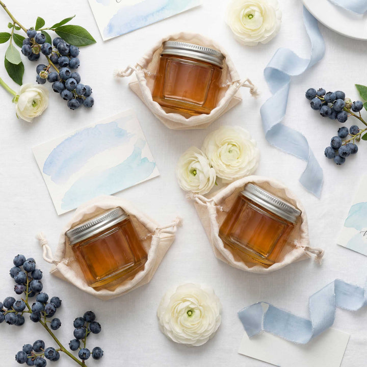 Three jars of honey on a white surface with flowers and blueberries.