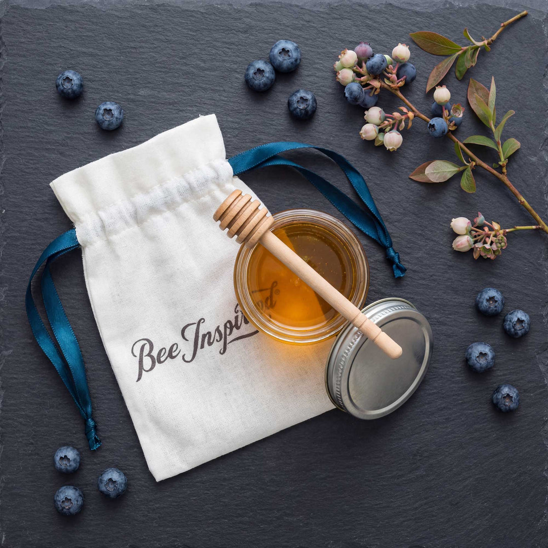 Honey jar with dipper, white pouch labeled 'Bee Inspired', and blueberries on a dark surface