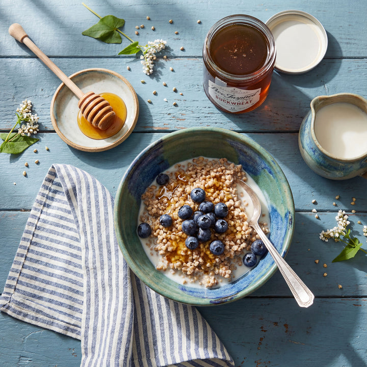 Bowl of cereal with blueberries, jar of honey, and pitcher on a blue wooden surface.