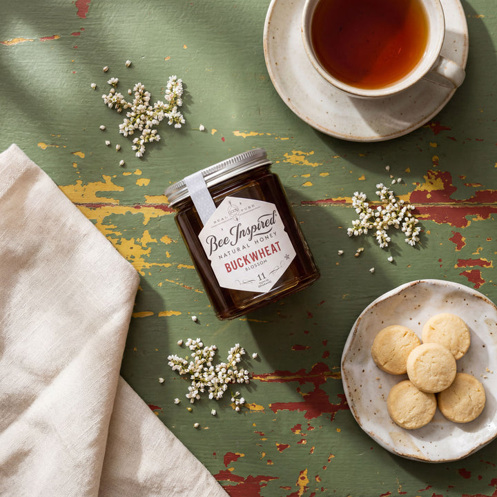 Jar of Bee Inspired Buckwheat honey on a rustic wooden table with tea and cookies.