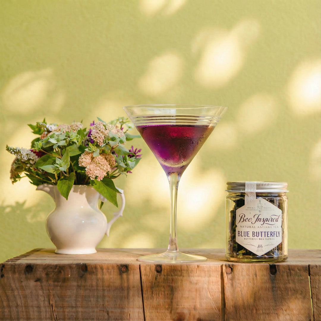 Purple cocktail in a martini glass with a jar of 'Blue Butterfly' and a vase of flowers on a wooden surface against a green wall.