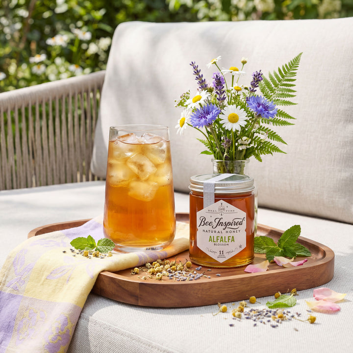 Bee-Inspired Alfalfa honey jar with iced tea and flowers on a wooden tray outdoors.