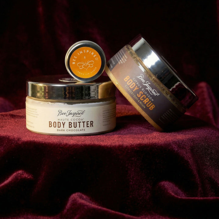 Container of body butter and scrub on a dark red fabric background topped with lemon lip gloss

